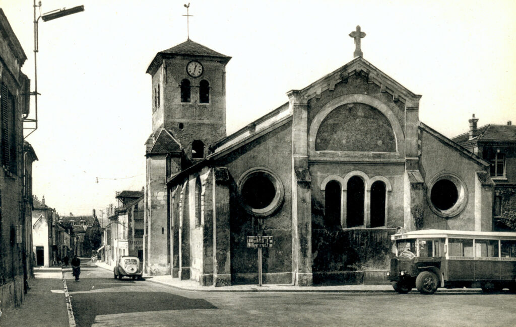 photo en noir et blanc de l’église en angle de rue goudronnée possédant un clocher surmonté d’une horloge et d’une girouette, des fenêtres arrondies - Agrandir l'image, fenêtre modale