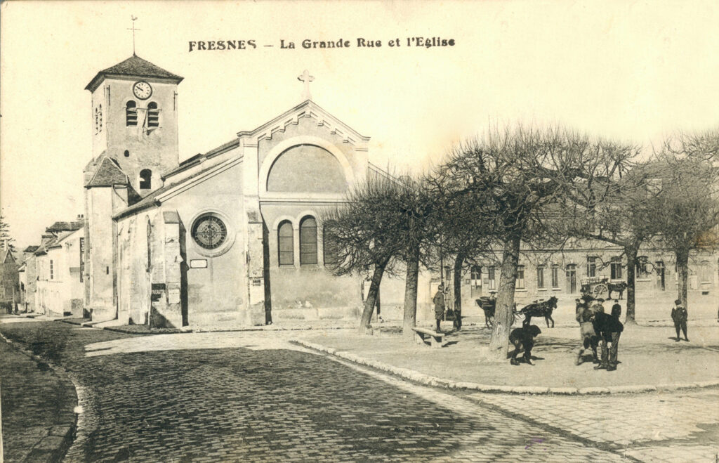 photo en noir et blanc de l’église sur la rue pavée, en face d’une placette entourée d’arbres, et des gens qui jouent, il est écrit « Fresnes - La Grande Rue et l’Église - Agrandir l'image, fenêtre modale