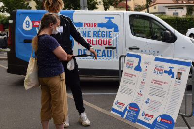 un homme avec un cahier et un stylo commente un panneau explicatif de la régie publique de l'eau à une femme - Agrandir l'image 1 sur 9, fenêtre modale